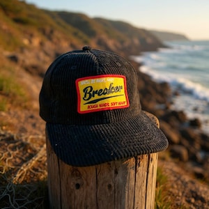 Pode incluir: Boné de beisebol de veludo cotelê preto com um patch amarelo e vermelho. O patch diz "Breaker" e as frases "Shell be right, mate" e "Rough hands, soft heart". O boné está em um poste de madeira.