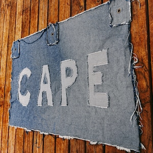 May include: A rectangular denim fabric piece with frayed edges, featuring the word CAPE appliqued in white and blue striped fabric. The denim has three reinforced metal grommets along the top edge for hanging or attachment purposes.