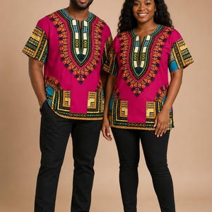 May include: Two matching short-sleeved shirts in vibrant magenta fabric featuring a central V-neckline and hemline decorated with intricate geometric patterns in yellow, blue, green, and black. Each shirt includes two front pockets and black pants.