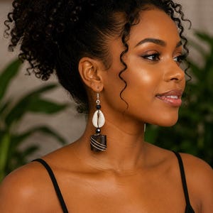 May include: A dangling earring featuring a silver-tone hook, a small black bead, a white cowrie shell, and a cylindrical dark brown wooden bead wrapped with thin silver wire. The earring hangs vertically with a layered, multi-material design.