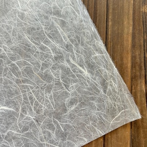May include: A sheet of handmade paper with a gray background and white, fibrous inclusions. The paper is angled, revealing a corner, and rests on a wooden surface with vertical grain.