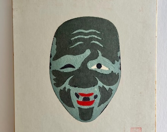 Könnte beinhalten: Vintage japanischer Noh-Theatermasken-Druck. Die Maske hat ein zwinkerndes Auge, einen lächelnden Mund mit roten Lippen und ein teal-schwarzes Farbschema. Der Druck befindet sich auf einem hellbeigen Hintergrund.