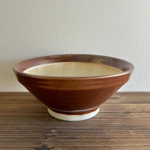 May include: A brown ceramic mortar and pestle bowl with a white interior and a white base. The bowl has a wide, shallow shape with a textured interior for grinding ingredients. The exterior is a glossy brown.