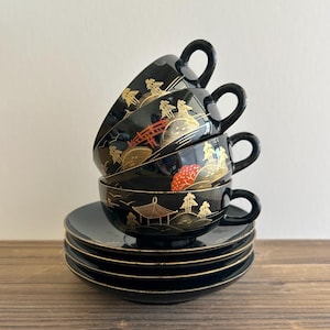May include: A set of four black teacups and saucers with gold and red painted Japanese-inspired designs. The cups and saucers are stacked on top of each other.