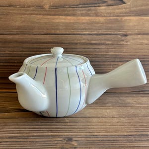 May include: White ceramic teapot with a lid and a side handle. The teapot is decorated with vertical stripes in red, blue, and green. The teapot is sitting on a wooden surface.