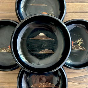 May include: A collection of six black lacquerware plates. Each plate features a gold landscape design, including mountains, trees, and water. The plates have a glossy finish and a round shape, perfect for serving appetizers or small dishes.