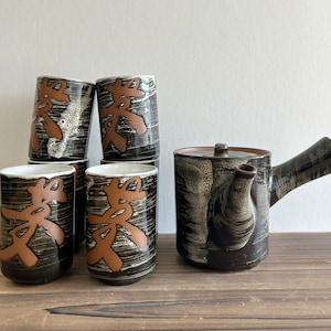 Könnte beinhalten: Ein Set aus sechs schwarzen und weißen Keramik-Teetassen und einer passenden Teekanne mit brauner Glasur und japanischen Schriftzeichen. Die Tassen und die Teekanne haben ein strukturiertes, pinselstrichähnliches Design.