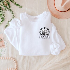 May include: A white sweatshirt featuring a black and white crest with a cross, book, and shield design, encircled by a laurel wreath. The text "TOMMEN COLLEGE" is displayed below the crest. A beige hat and decorative greenery are in the background.