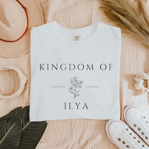 May include: White t-shirt with a black graphic design that says "Kingdom of" with a floral illustration and the text "Hunted" and "Hunter" on either side of the flower. Below the flower is the name "Ilya".