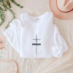 Puede incluir: Sudadera blanca con un gráfico negro de una espada y la palabra "POWERLESS" impresa en la parte delantera.