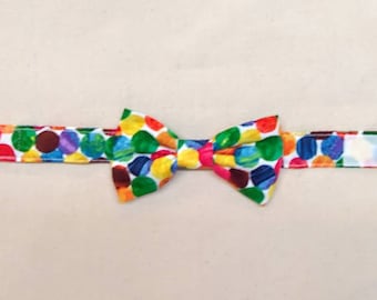 May include: A bow tie featuring a vibrant, multi-colored polka dot pattern in shades of red, yellow, green, blue, and purple. The bow tie is attached to a matching adjustable fabric strap that secures with a white hook and loop fastener.