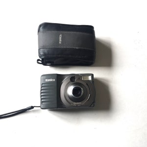 May include: A black Konica Q-Maxov camera with a black leather case. The camera has a lens cover and a strap.