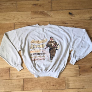 Puede incluir: Una sudadera gris claro con un gráfico vintage de un corredor que lleva una bolsa. El gráfico incluye el texto "Sporting Club Champ" y "The Best Competition for His and Her Training Forced".