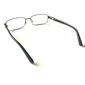 May include: Black metal frame eyeglasses with clear lenses. The glasses are shown from the back, with the arms extending out to the sides.