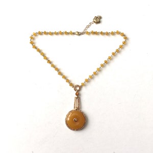 Puede incluir: Un collar de oro con un colgante redondo amarillo. El colgante está rodeado de pequeños cristales transparentes. El collar está hecho de pequeñas cuentas amarillas.