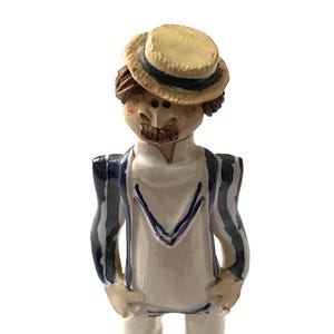 Puede incluir: Figura de cerámica de un hombre con sombrero de paja con banda negra, camisa blanca y chaqueta a rayas azules. La figura tiene bigote y está de pie con las manos en los bolsillos.