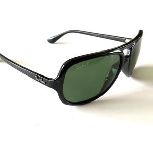 May include: Black Ray-Ban sunglasses with green lenses. The sunglasses have a thick, glossy black frame and the Ray-Ban logo is visible on the temple and lens. The lenses are a dark green color.