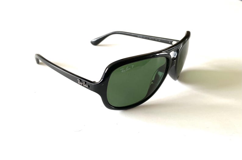 May include: Black Ray-Ban sunglasses with green lenses. The sunglasses have a thick, glossy black frame and the Ray-Ban logo is visible on the temple and lens. The lenses are a dark green color.