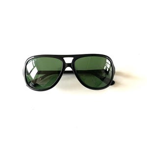 May include: Black aviator sunglasses with green lenses. The sunglasses have a thick black frame and a double bridge. The lenses are a dark green color, and the sunglasses are set against a white background.