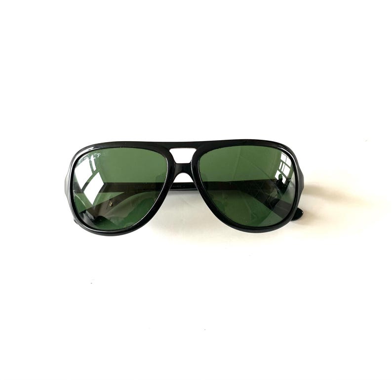 May include: Black aviator sunglasses with green lenses. The sunglasses have a thick black frame and a double bridge. The lenses are a dark green color, and the sunglasses are set against a white background.