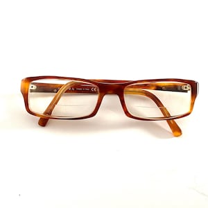 May include: Rectangular eyeglasses with a tortoiseshell pattern. The frames are brown and amber, with clear lenses. The Prada logo is visible on the frame. The glasses are set against a white background.