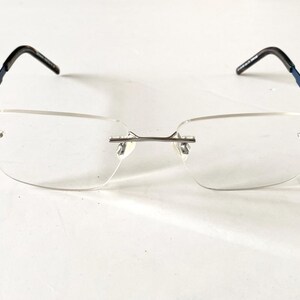 May include: Rimless eyeglasses with rectangular lenses. The temples are navy blue with black ear pieces. The bridge and nose pads are silver. The glasses are set against a white background.
