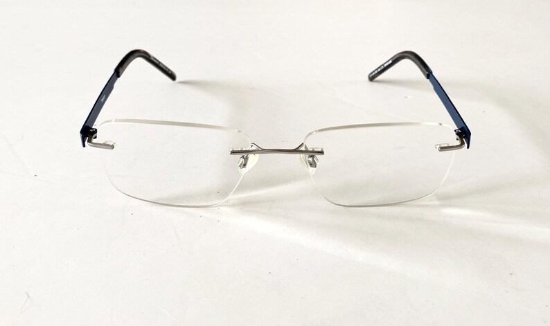 May include: Rimless eyeglasses with rectangular lenses. The temples are navy blue with black ear pieces. The bridge and nose pads are silver. The glasses are set against a white background.
