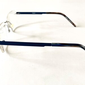 May include: Rimless eyeglasses with a navy blue frame and temples with a marbled brown and blue pattern. The lenses are clear, and the glasses are set against a white background. The word "Titanium" is printed on the frame.