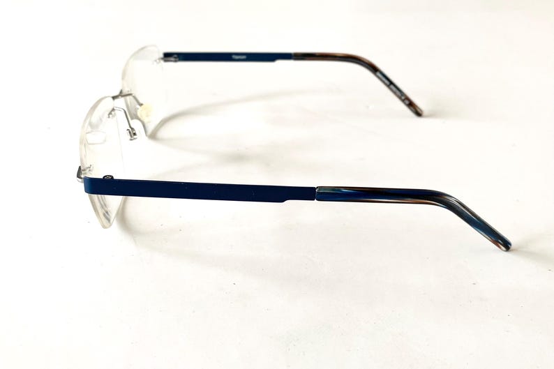 May include: Rimless eyeglasses with a navy blue frame and temples with a marbled brown and blue pattern. The lenses are clear, and the glasses are set against a white background. The word "Titanium" is printed on the frame.