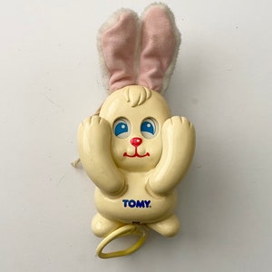 May include: A white plastic wind-up toy bunny with pink fabric ears. The bunny has blue eyes, a red nose, and a yellow pull string. The toy is labeled "TOMY."