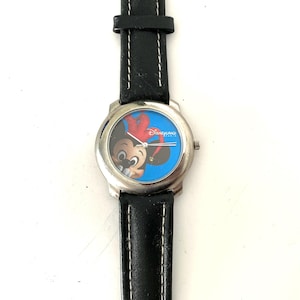 Puede incluir: Un reloj de pulsera plateado con una correa de cuero negro. La esfera del reloj presenta a Mickey Mouse sobre un fondo azul, con la palabra "Disneyland" en rojo. La correa está cosida con hilo blanco.