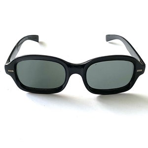 Może przedstawiać: Czarne okulary przeciwsłoneczne z tworzywa sztucznego z ciemnoszarymi szkłami. Okulary mają zaokrąglony prostokątny kształt.