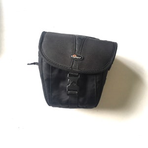 May include: Black camera bag with a zipper and a buckle closure. The bag has a small orange logo that says "Lowepro" on the front.