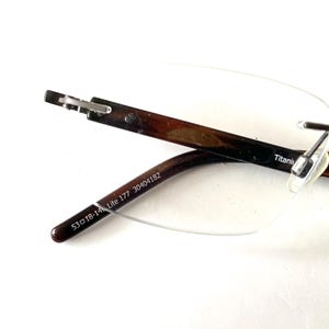 Peut inclure: Lunettes sans monture avec des branches &agrave; motifs marron et noir. Les branches portent l'inscription "53 18-14 Lite 177 30404182". La monture est en titane. Les verres sont transparents.