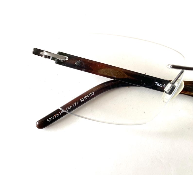 Peut inclure: Lunettes sans monture avec des branches &agrave; motifs marron et noir. Les branches portent l'inscription "53 18-14 Lite 177 30404182". La monture est en titane. Les verres sont transparents.