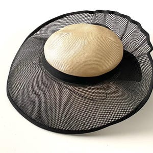 Può includere: Un cappello di rete nero a larghe falde con una corona di paglia beige. Il cappello ha un bordo arricciato e un nastro nero attorno alla corona.