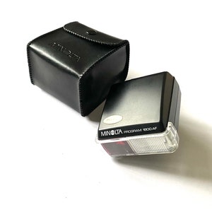 以下が含まれることがあります： 白いディフューザーと赤いフラッシュバルブが付いた黒いミノルタプログラム1800 AFフラッシュユニット。フラッシュユニットは黒い革ケースに入っています。