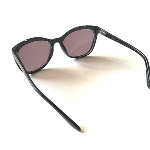 Pode incluir: &Oacute;culos de sol pretos de olho de gato com lentes escuras. Os &oacute;culos s&atilde;o de pl&aacute;stico e t&ecirc;m um design elegante e moderno.