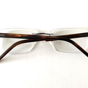 Peut inclure: Lunettes sans monture avec des branches et un pont marron et &eacute;caille de tortue. Les verres sont clairs et rectangulaires. Les lunettes sont pos&eacute;es sur un fond blanc.