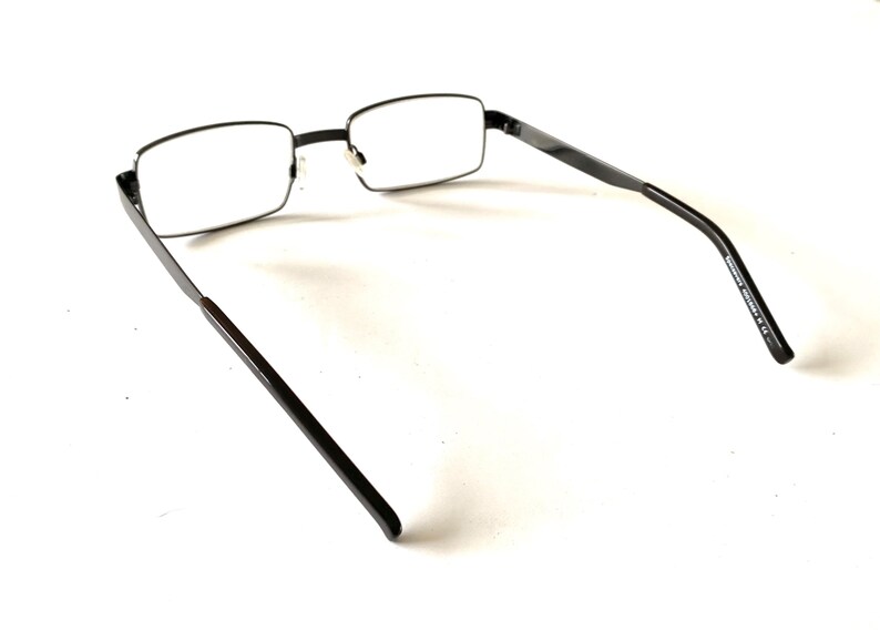 May include: Black rectangular eyeglasses with clear lenses. The frames are a dark, matte finish, and the temples have a slight curve. The glasses are set against a white background.
