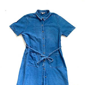 Könnte beinhalten: Kurzärmeliges Jeanskleid in mittlerer Blaufärbung. Das Kleid hat einen Kragen, eine Knopfleiste vorne, zwei Vordertaschen und einen passenden Gürtel, der in der Taille gebunden ist. Das Kleid ist aus leichtem Jeansstoff gefertigt.