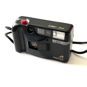 Peut inclure: Appareil photo compact Pentax PC-303 noir avec un bouton de déclenchement rouge et une dragonne noire. L'appareil photo dispose d'un flash intégré et le logo Pentax est visible à l'avant.