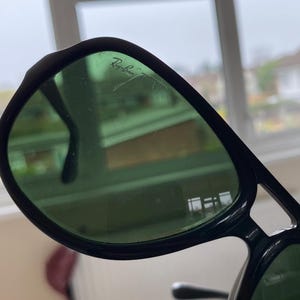 May include: Close-up of black Ray-Ban sunglasses with green tinted lenses. The Ray-Ban logo is visible on the upper left lens. The sunglasses have a classic aviator style with a thick frame.