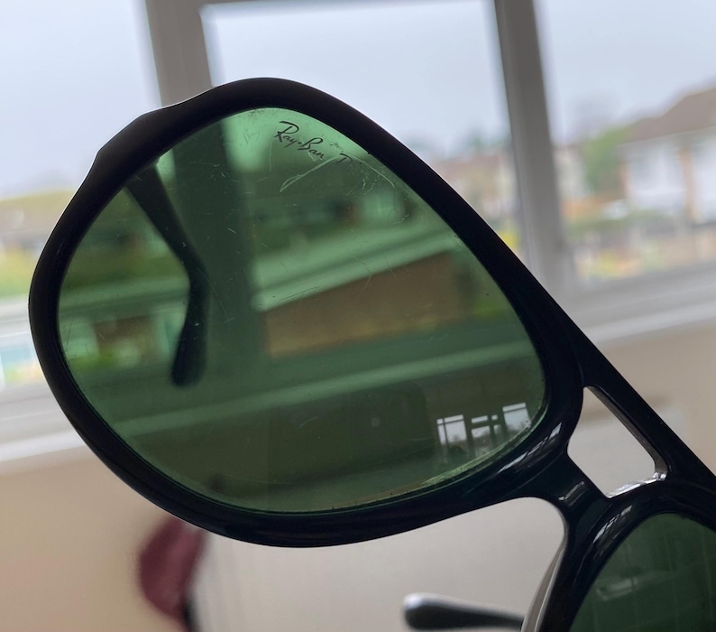 May include: Close-up of black Ray-Ban sunglasses with green tinted lenses. The Ray-Ban logo is visible on the upper left lens. The sunglasses have a classic aviator style with a thick frame.