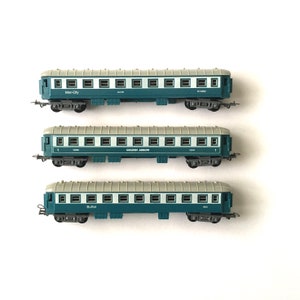 Puede incluir: Tres vagones de tren en miniatura, cada uno con un nombre diferente: Inter-City, Golden Arrow y Buffet. Los vagones son azules y grises con ventanas blancas y están dispuestos en una pila vertical.