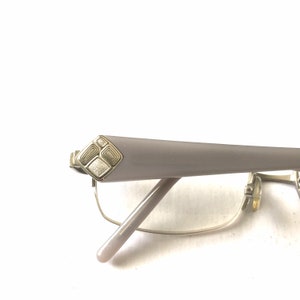 May include: A pair of silver-toned eyeglasses with a light gray frame. The frame has a decorative design on the temple.