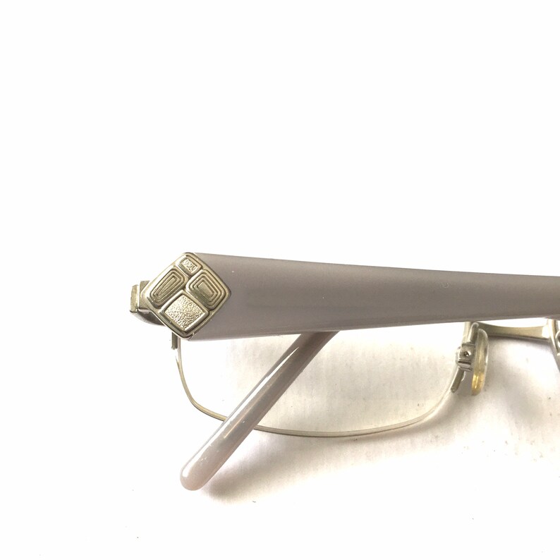 May include: A pair of silver-toned eyeglasses with a light gray frame. The frame has a decorative design on the temple.