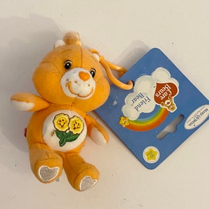 Może przedstawiać: Żółty pluszowy brelok Care Bears z kwiatowym wzorem na klatce piersiowej. Brelok jest przymocowany do niebiesko-białej karty z napisem "Care Bears Friend Bear".