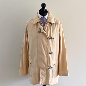 Può includere: Una giacca giallo chiaro con colletto ripiegato, maniche lunghe e chiusura frontale con quattro alamari in metallo. La giacca presenta due tasche anteriori inferiori con patta ed è esposta sopra una camicia bianca con cravatta blu.