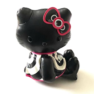 May include: Black Hello Kitty figurine with a pink bow and accents. The figurine has a black body with a white and black patterned dress. The bow has a pink outline and white and black details. The figurine is sitting down.